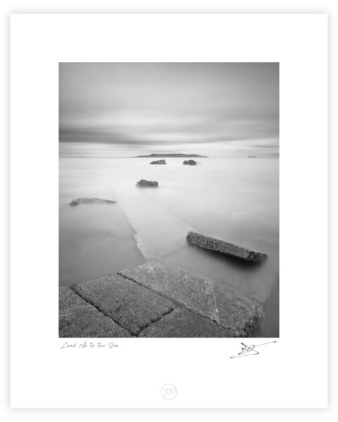 Black and white long exposure from Sandycove slipway looking across Dublin Bay toward Howth Head, with stone steps leading into calm water.
