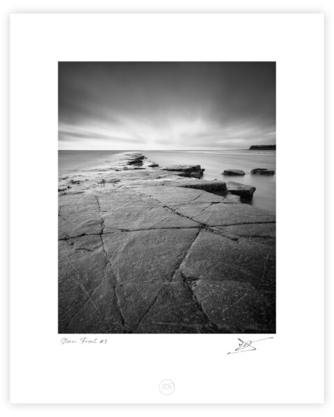 Rock shelf and calm sea beneath storm clouds at Kimmeridge Bay on the Dorset coast, black and white photograph.