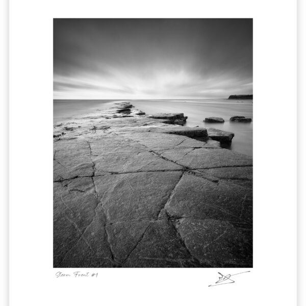 Rock shelf and calm sea beneath storm clouds at Kimmeridge Bay on the Dorset coast, black and white photograph.