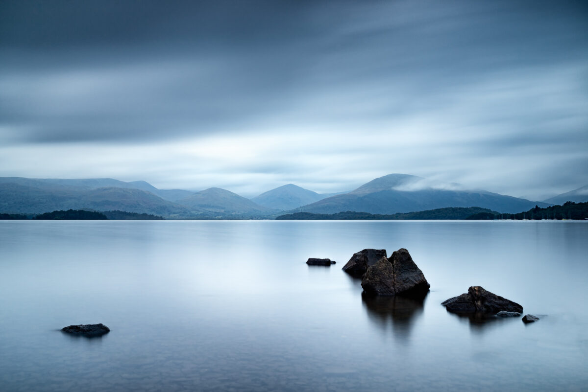 Come Sit with Me Awhile Stones in calm water at Millarochy Bay on Loch Lomond with mountains and fast-moving clouds, Scotland, colour photograph.
