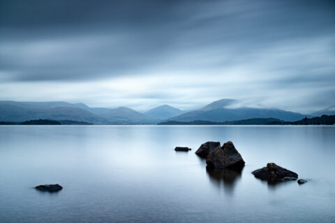 Stones in calm water at Millarochy Bay on Loch Lomond with mountains and fast-moving clouds, Scotland, colour photograph.