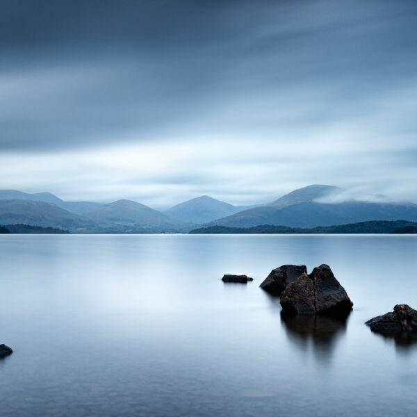 Stones in calm water at Millarochy Bay on Loch Lomond with mountains and fast-moving clouds, Scotland, colour photograph.