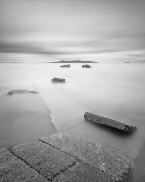 Black and white long exposure from Sandycove slipway looking across Dublin Bay toward Howth Head, with stone steps leading into calm water.
