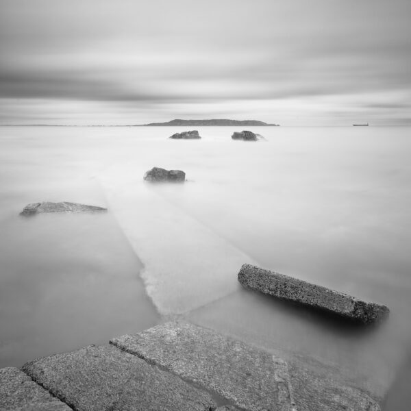 Black and white long exposure from Sandycove slipway looking across Dublin Bay toward Howth Head, with stone steps leading into calm water.