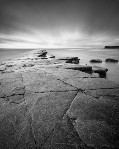 Rock shelf and calm sea beneath storm clouds at Kimmeridge Bay on the Dorset coast, black and white photograph.