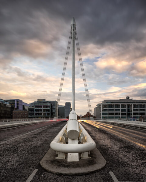 Samuel Beckett Bridge in Dublin seen head-on at dusk with long-exposure car light trails and dramatic clouds.