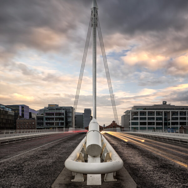 Samuel Beckett Bridge in Dublin seen head-on at dusk with long-exposure car light trails and dramatic clouds.
