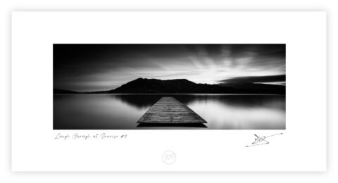 Black and white image of a wooden jetty extending into still water on Lough Caragh with mountains silhouetted behind.