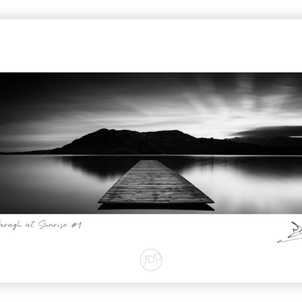 Black and white image of a wooden jetty extending into still water on Lough Caragh with mountains silhouetted behind.