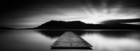 Black and white image of a wooden jetty extending into still water on Lough Caragh with mountains silhouetted behind.
