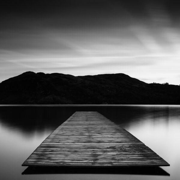 Black and white image of a wooden jetty extending into still water on Lough Caragh with mountains silhouetted behind.