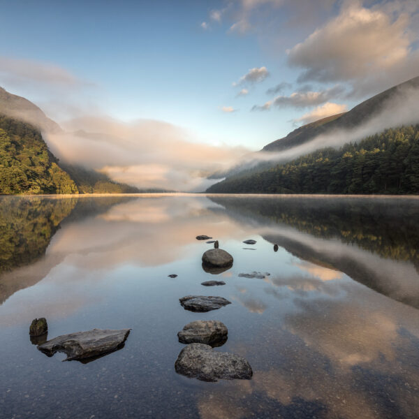 Mist rising over Upper Lake in Glendalough with rocks reflected in still water in County Wicklow.