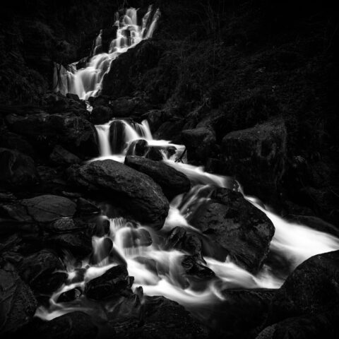 Black and white photograph of Torc Waterfall flowing over rocks in Killarney National Park, Ireland.