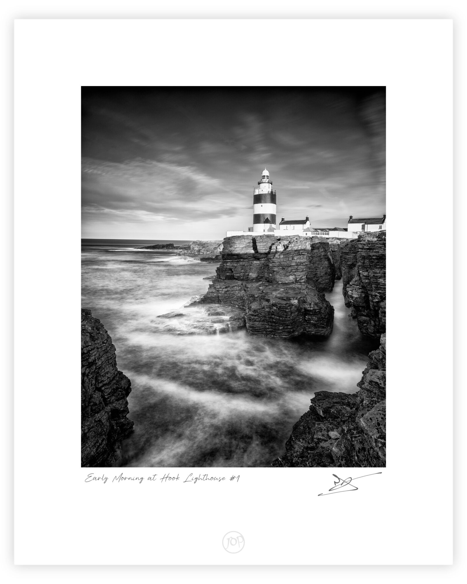 Black and white photograph of Hook Lighthouse above rocky cliffs with waves moving around the shoreline in County Wexford, Ireland.