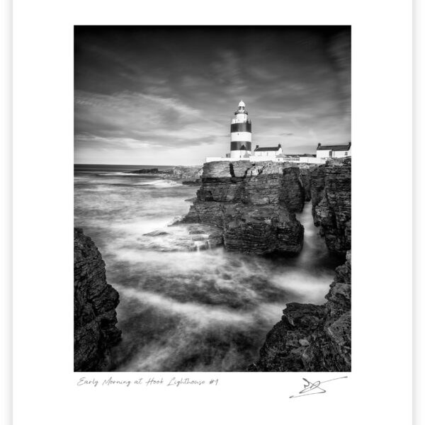 Black and white photograph of Hook Lighthouse above rocky cliffs with waves moving around the shoreline in County Wexford, Ireland.