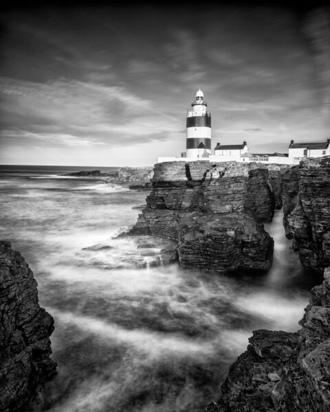 Black and white photograph of Hook Lighthouse above rocky cliffs with waves moving around the shoreline in County Wexford, Ireland.