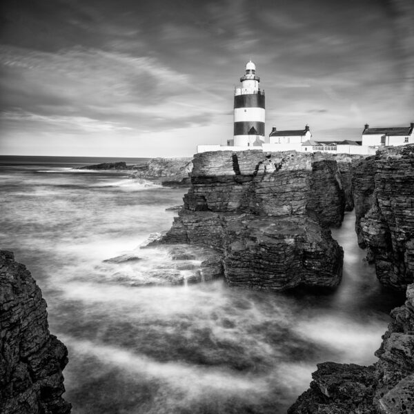 Black and white photograph of Hook Lighthouse above rocky cliffs with waves moving around the shoreline in County Wexford, Ireland.