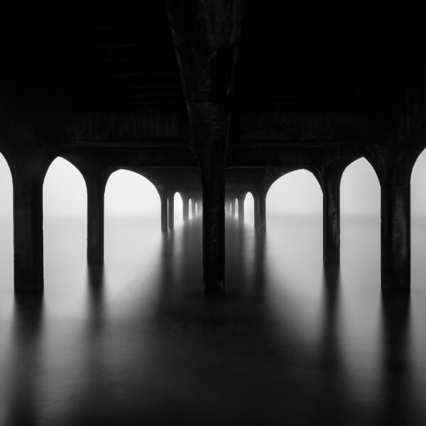 Under Boscombe Pier at dawn with still water and repeating arches in Bournemouth, England, black and white photograph.