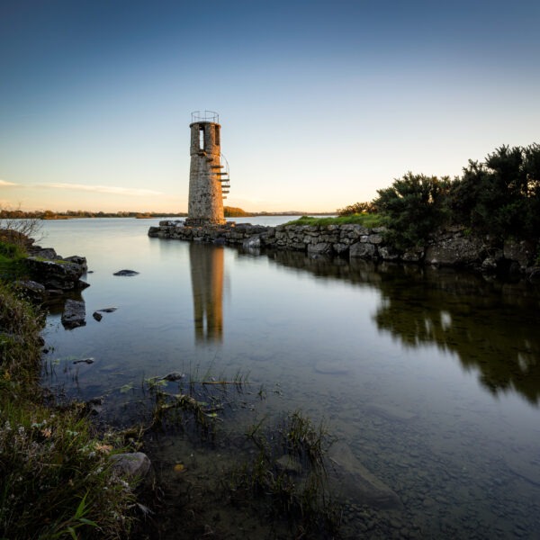 Ballycurrin Lighthouse reflected in calm water on Lough Corrib at sunset in Galway, Ireland, colour photograph.