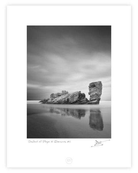 Black and white long exposure photograph of an elongated sea stack with a taller upright pillar beside it, reflected in wet sand at Playa de Requesinos, Asturias, Spain.