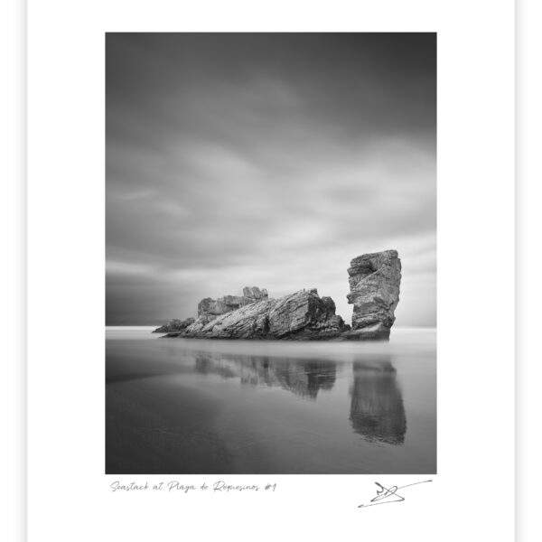 Black and white long exposure photograph of an elongated sea stack with a taller upright pillar beside it, reflected in wet sand at Playa de Requesinos, Asturias, Spain.