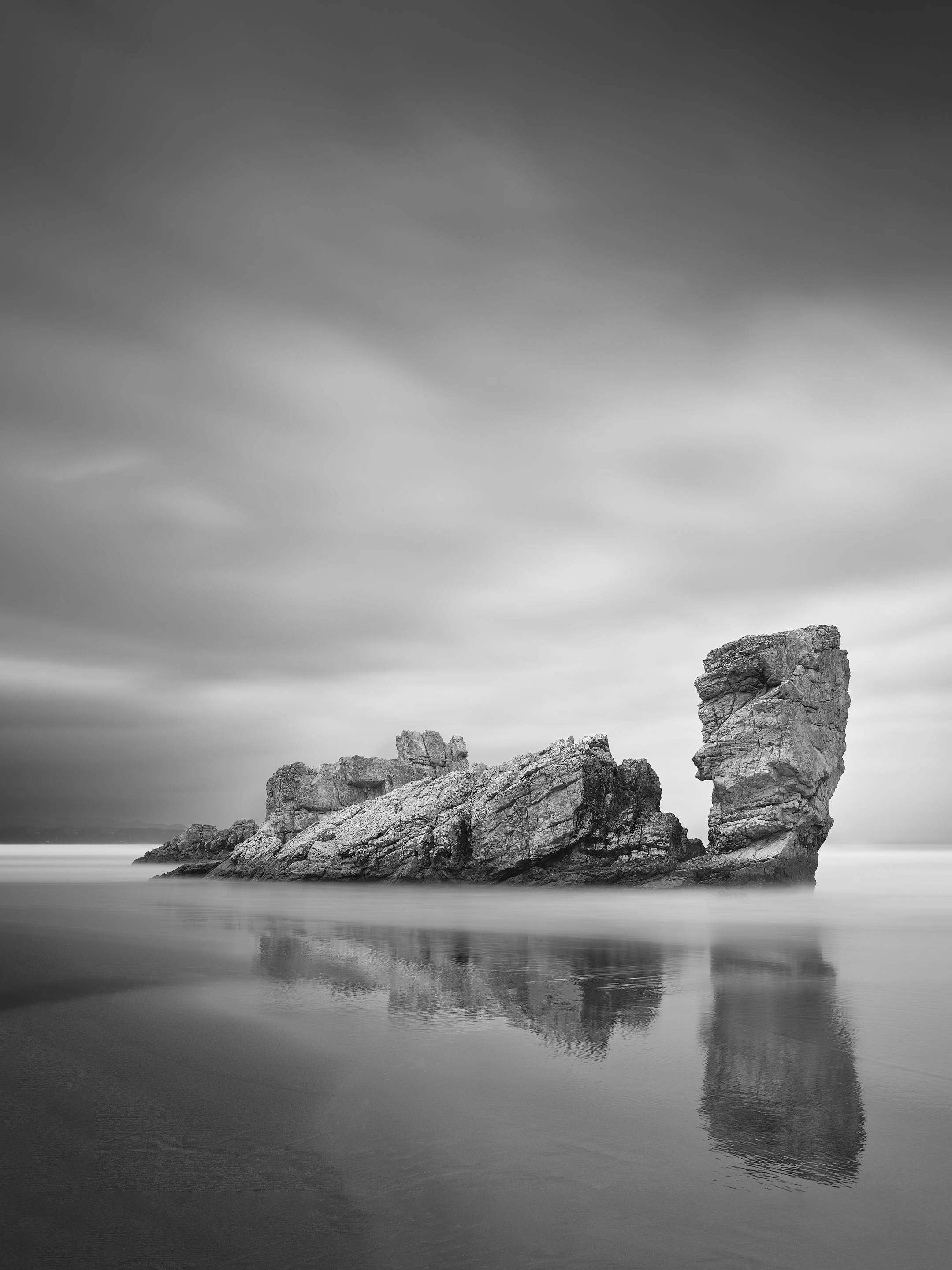 Black and white long exposure photograph of an elongated sea stack with a taller upright pillar beside it, reflected in wet sand at Playa de Requesinos, Asturias, Spain.