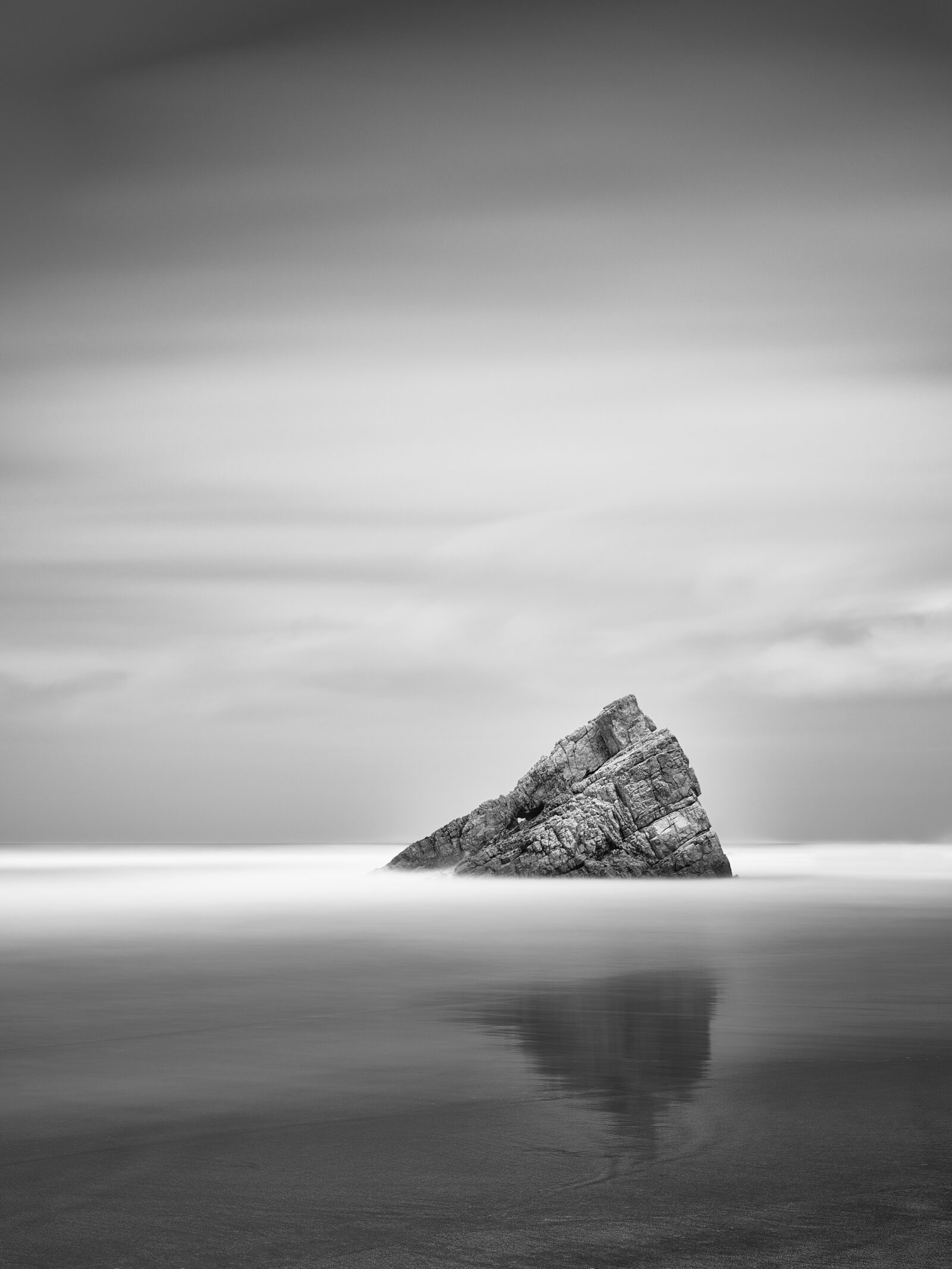 Black and white long exposure photograph of a leaning sea stack rising from calm reflective water at Playa de Requesinos, Asturias, Spain.