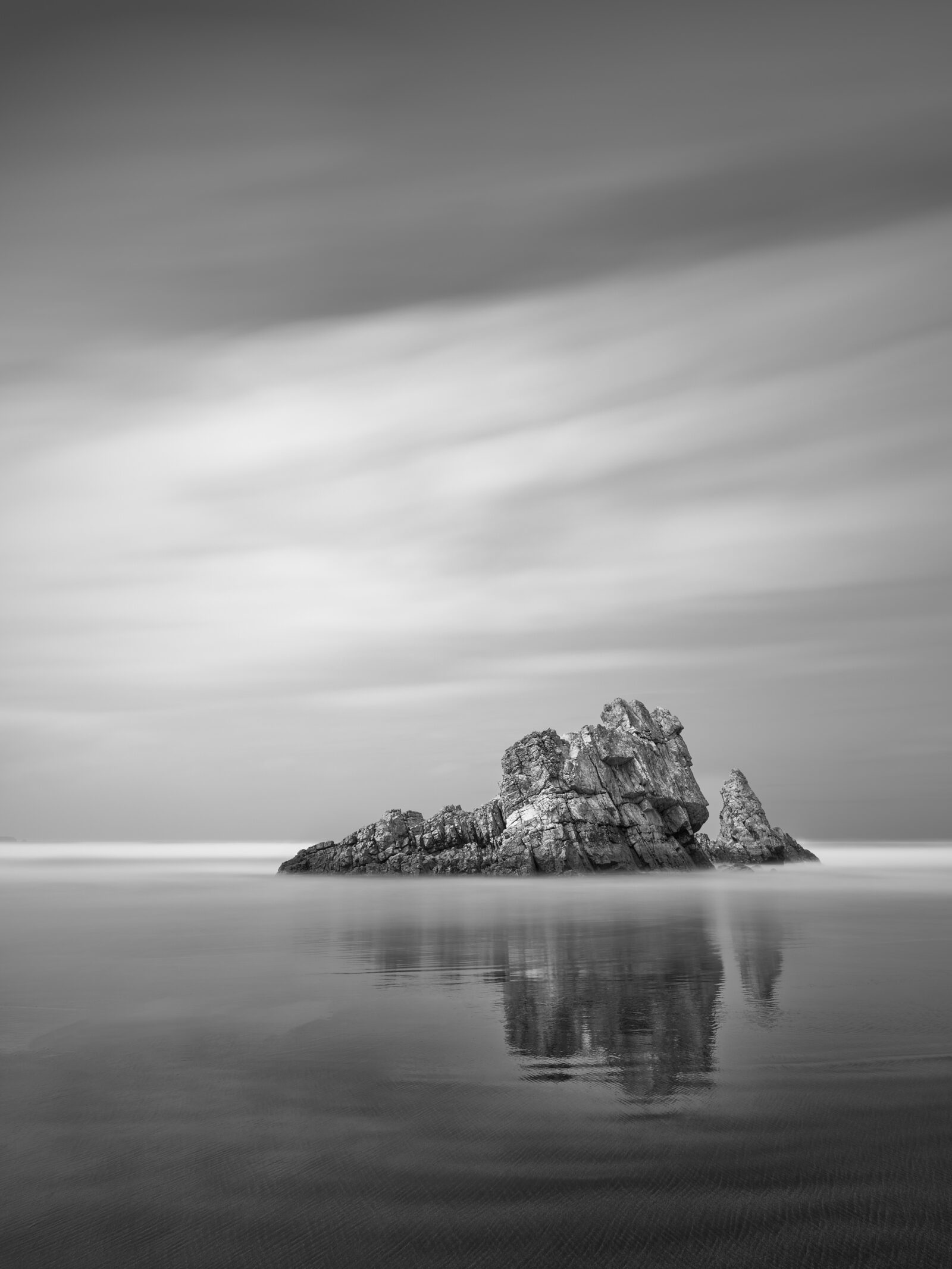 Black and white long exposure photograph of a low, jagged sea stack emerging from smooth tidal water with a soft sky above, Playa de Requesinos, Asturias, Spain.