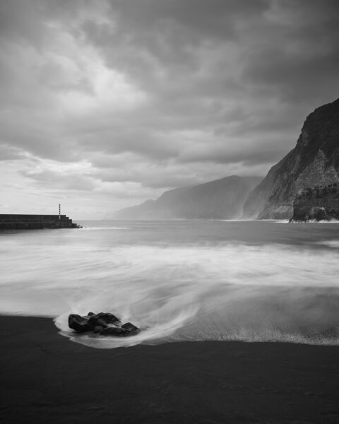 Black-and-white vertical view of waves sweeping around a dark rock on the black-sand beach at Praia do Seixal, with a harbour wall and striped beacon at left and steep cliffs receding along the coast at right.