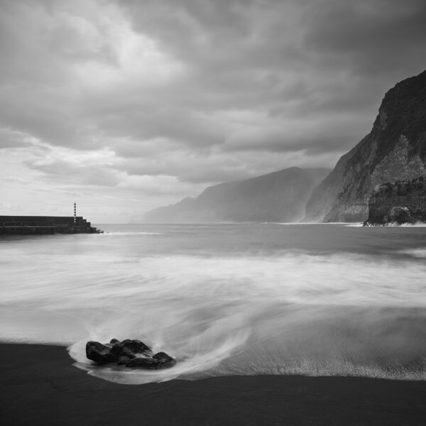 Black-and-white vertical view of waves sweeping around a dark rock on the black-sand beach at Praia do Seixal, with a harbour wall and striped beacon at left and steep cliffs receding along the coast at right.