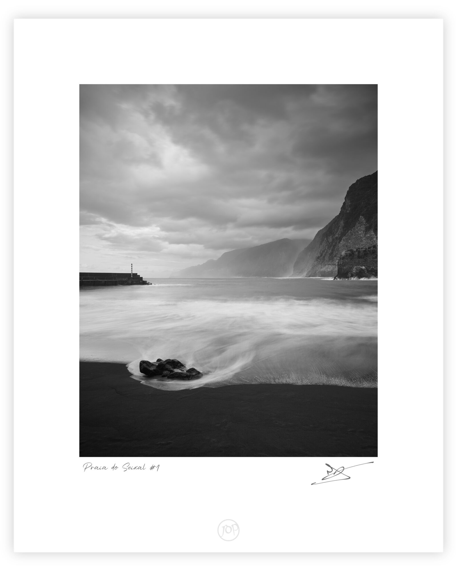 Black-and-white vertical view of waves sweeping around a dark rock on the black-sand beach at Praia do Seixal, with a harbour wall and striped beacon at left and steep cliffs receding along the coast at right.