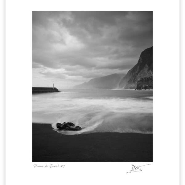 Black-and-white vertical view of waves sweeping around a dark rock on the black-sand beach at Praia do Seixal, with a harbour wall and striped beacon at left and steep cliffs receding along the coast at right.
