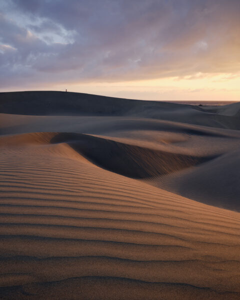 Layered sand dunes at Maspalomas curve across the foreground and middle distance, their rippled surfaces lit by warm sunrise light beneath heavy blue-grey clouds, with a lone person standing on the far ridge and a narrow strip of sea beyond.