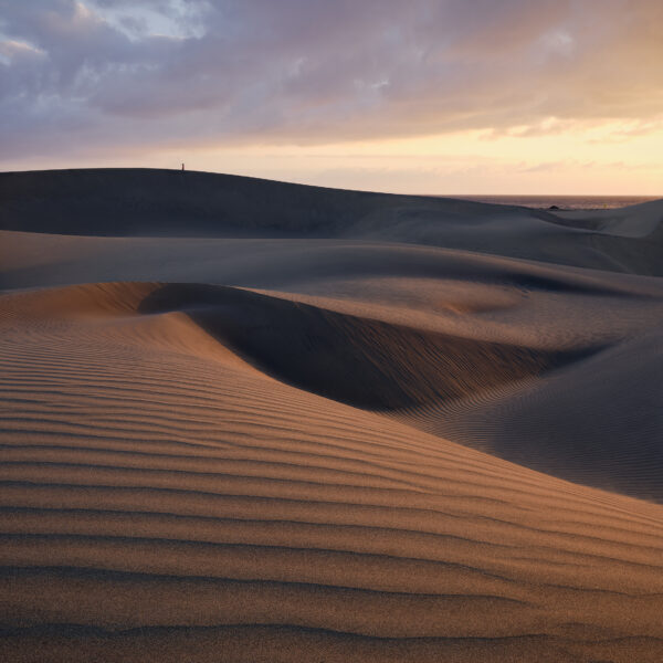 Layered sand dunes at Maspalomas curve across the foreground and middle distance, their rippled surfaces lit by warm sunrise light beneath heavy blue-grey clouds, with a lone person standing on the far ridge and a narrow strip of sea beyond.