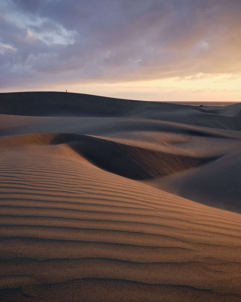 Layered sand dunes at Maspalomas curve across the foreground and middle distance, their rippled surfaces lit by warm sunrise light beneath heavy blue-grey clouds, with a lone person standing on the far ridge and a narrow strip of sea beyond.