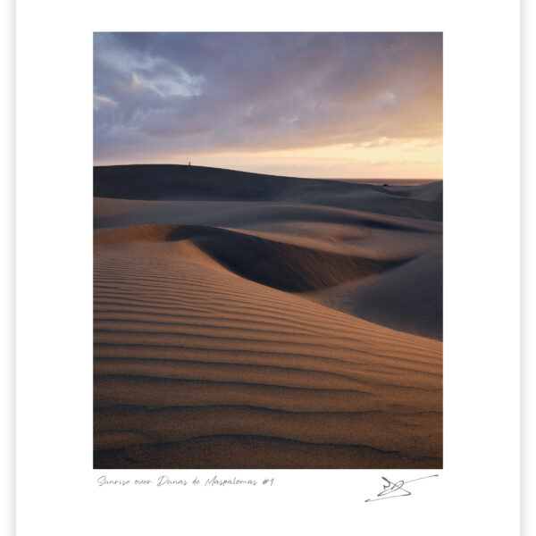 Layered sand dunes at Maspalomas curve across the foreground and middle distance, their rippled surfaces lit by warm sunrise light beneath heavy blue-grey clouds, with a lone person standing on the far ridge and a narrow strip of sea beyond.