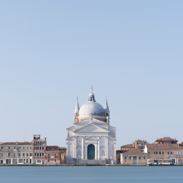 Centred view across smooth blue water to the white Basilica del Santissimo Redentore in Venice, framed by low terracotta-roofed buildings beneath a vast cloudless sky.