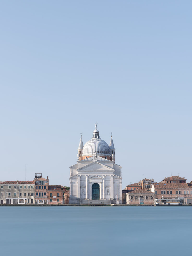 Centred view across smooth blue water to the white Basilica del Santissimo Redentore in Venice, framed by low terracotta-roofed buildings beneath a vast cloudless sky.