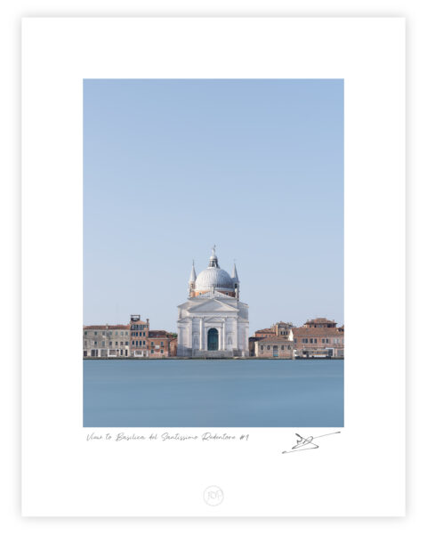 Centered view across smooth blue water to the white Basilica del Santissimo Redentore in Venice, framed by low terracotta-roofed buildings beneath a vast cloudless sky.