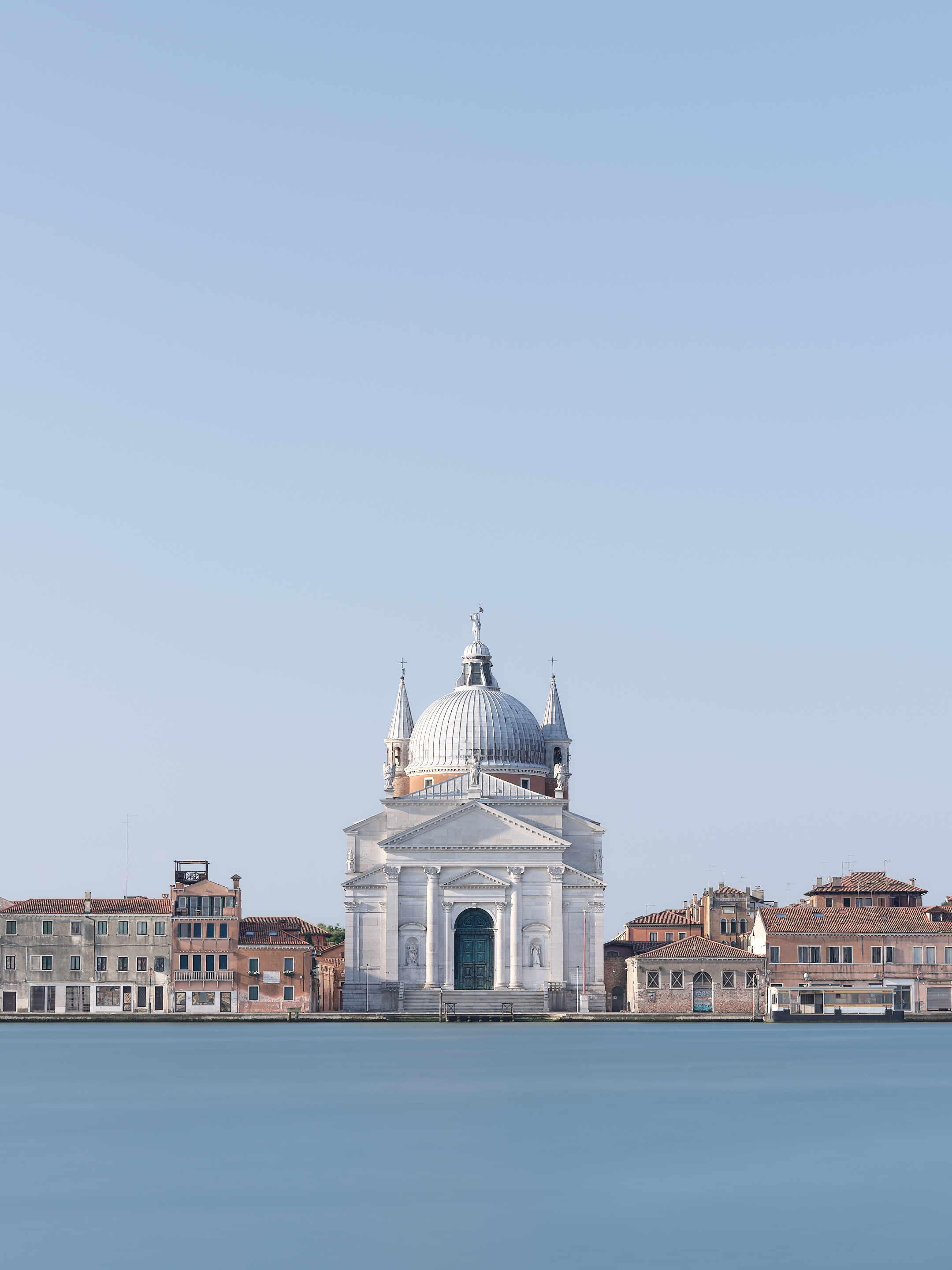 Centred view across smooth blue water to the white Basilica del Santissimo Redentore in Venice, framed by low terracotta-roofed buildings beneath a vast cloudless sky.