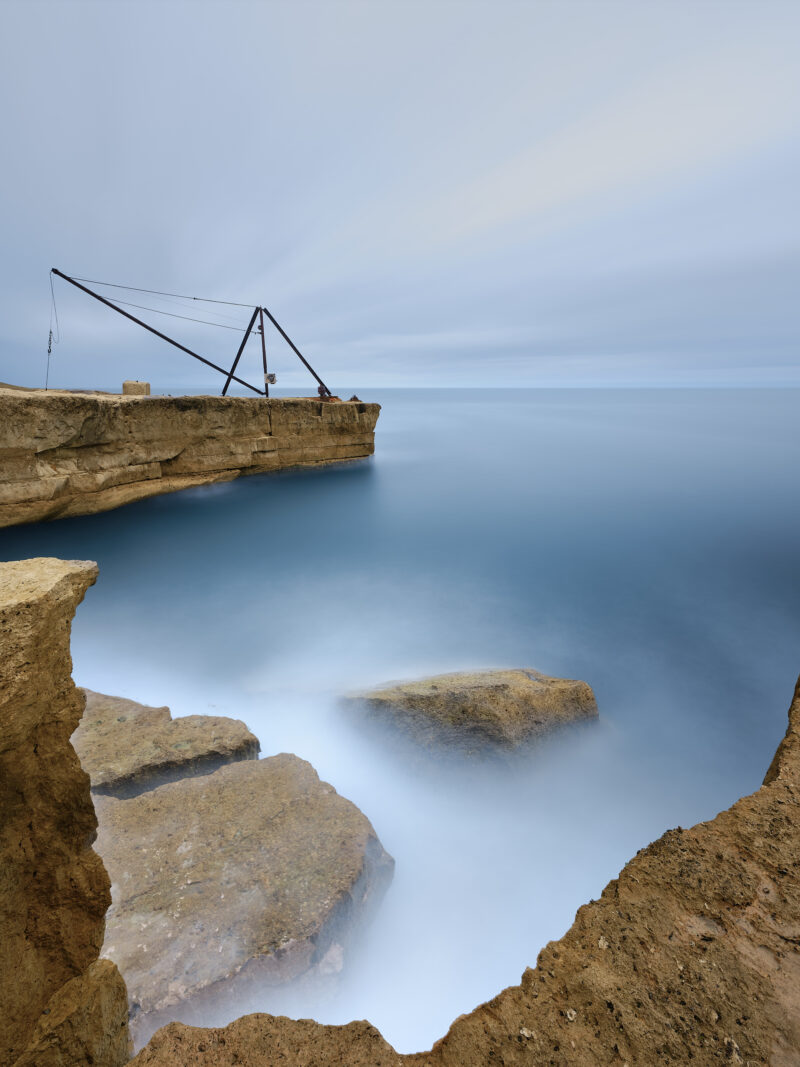 A dark metal crane stands on a rock ledge at upper left, with ochre rocks framing smooth blue water in the foreground and pale cloud above, at Portland Bill.